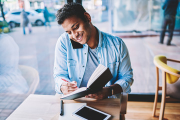 Handsome positive smart businessman communicating with friends via application on smartphone while writing down to notepad.Good looking smiling male person recording information and talking on phone