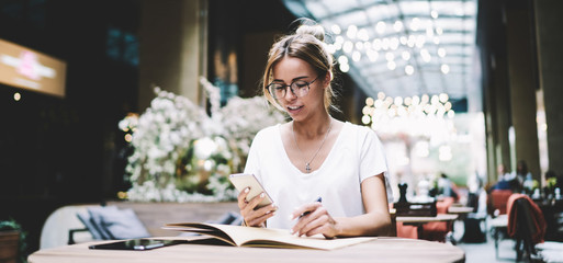 Caucasian female blogger reading messages from followers in social network while making research of information for creating new post, young woman in eyewear installing application on new cellphone