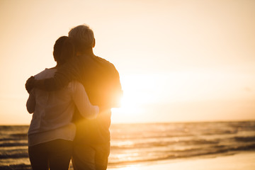 Fototapeta premium Rear view of elderly couple standing hugging on sandy beach during sunset, Retirement age concept.