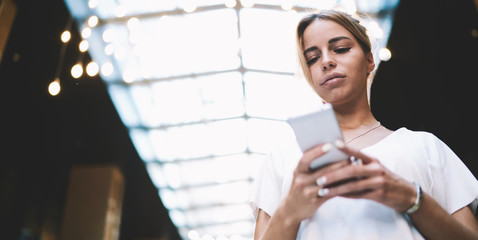 Attractive young hipster girl sending text message via smartphone gadget connected to 4g internet indoors, caucasian woman dressed in casual look watching video online via digital cellular phone