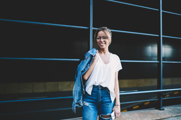 Fototapeta premium Half length portrait of cheerful hipster girl in optical eyewear for vision correction looking at camera during leisure time on publicity area, happy female teenager in denim apparel outdoors