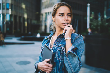 Fashionable woman dreaming about future vacations while spending time on publicity area with modern touchpad in hands, charming hipster girl in jeans jacket holding digital tablet and looking away