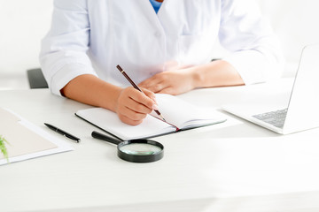 cropped view of dermatologist sitting at table and writing in notebook in clinic