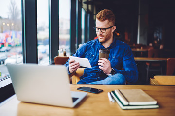 Obraz premium Male freelancer in eyeglasses watching video online via modern touch pad during break from working on laptop, millennial man enjoying coffee break during sharing files via digital tablet and netbook