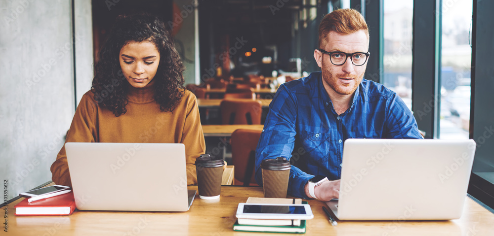Poster portrait of serious hipster guy looking at camera while female colleague near typing program code fo