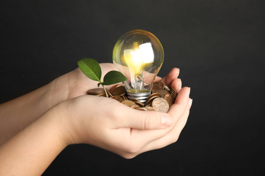 Woman with coins, light bulb and green plant against black background, closeup. Power saving
