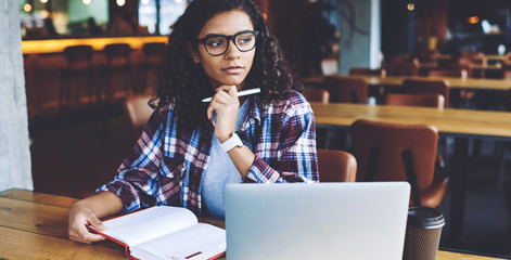 Pondering african female student thinking on school homework during learning in university campus, thoughtful woman in eyewear looking away while sitting at table with computer and knowledge textbook