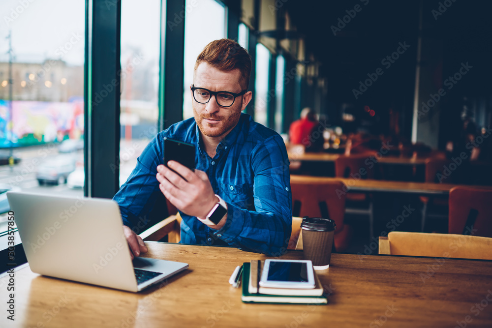 Poster skilled student with red hair reading incoming notification on smartphone device while working on co