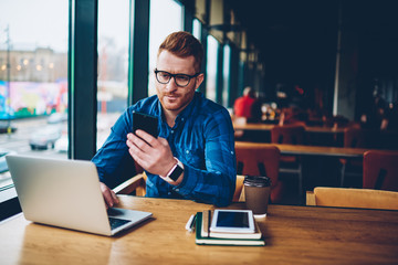 Skilled student with red hair reading incoming notification on smartphone device while working on...