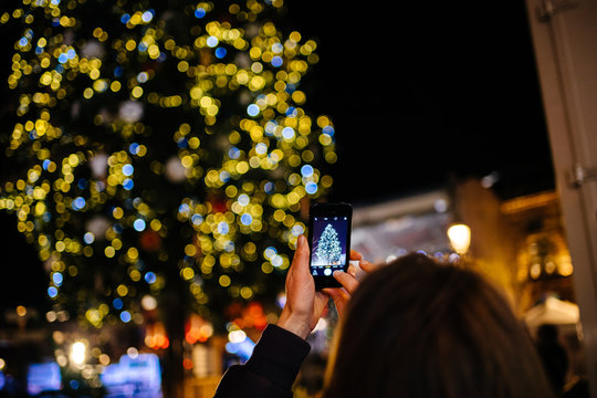 Strasbourg, France - Nov 23, 2017: Rear view of adult woman rising smartphone to take a photograph on smartphone the illuminated and decorated Christmas fir tree at the market capital