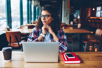 Thoughtful african american young woman looking out of window and thinking on new studying project and working plan sitting at digital computer connected to 4G internet in stylish coworking space