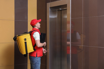 Male courier with thermo bag and clipboard waiting for elevator. Food delivery service