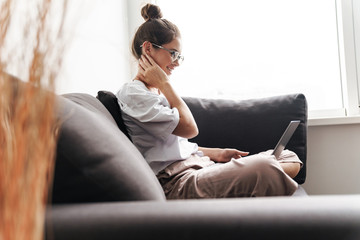 Image of pleased cute woman typing on laptop while sitting on sofa - obrazy, fototapety, plakaty