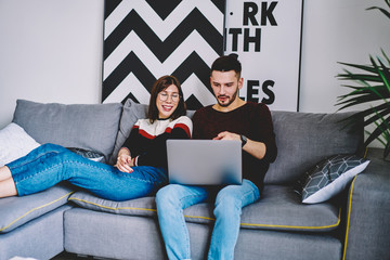 Caucasian man booking tickets on website via laptop sitting on comfortable couch with girlfriend using home internet connection, couple in love shopping together on computer while resting on sofa