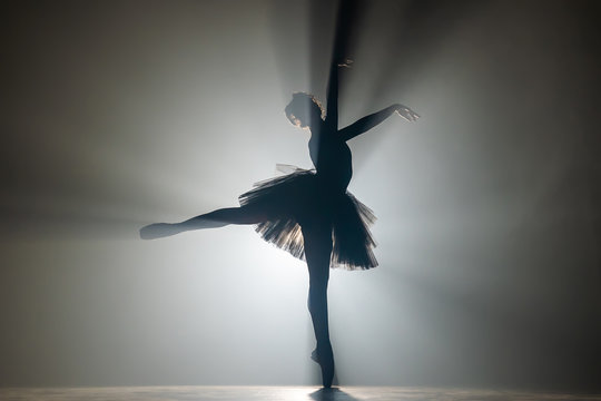 Fototapeta Professional ballerina dancing ballet in spotlights smoke on big stage. Beautiful young girl wearing black tutu dress on floodlights background. Black and white.