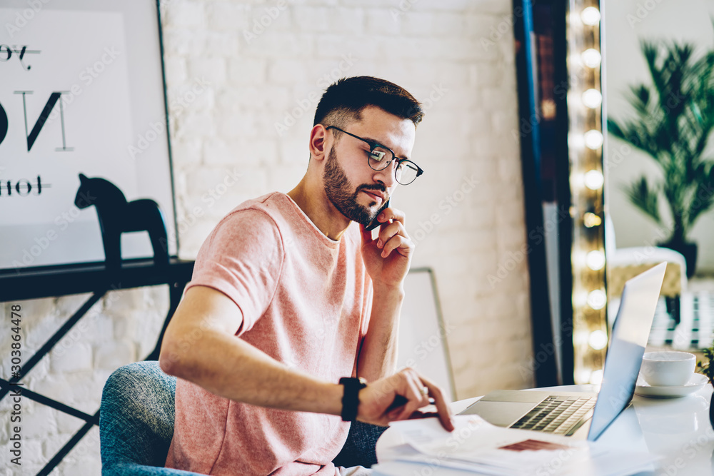 Poster young man calling on smartphone to operator and checking banking account sitting at modern laptop in