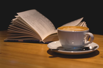 Ceramic cup of cappuccino in coffee shop with pattern on a wooden table with an open book. Latte...