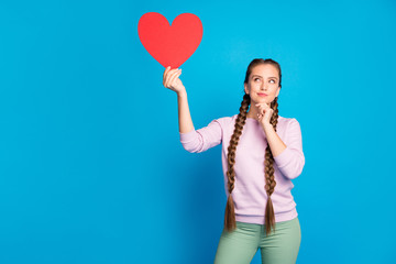 Portrait of thoughtful interested youth girl with long pigtails hold red paper card heart gift for valentine day think about cupid date wear pink pullover green pants isolated blue color background