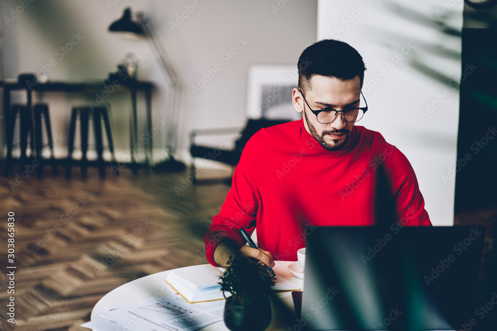 Poster concentrated bearded student studying at modern laptop computer and writing down notes for course wo