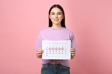 Young woman holding calendar with marked menstrual cycle days on pink background