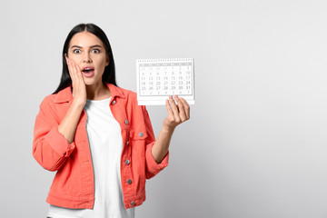 Emotional young woman holding calendar with marked menstrual cycle days on light background. Space for text