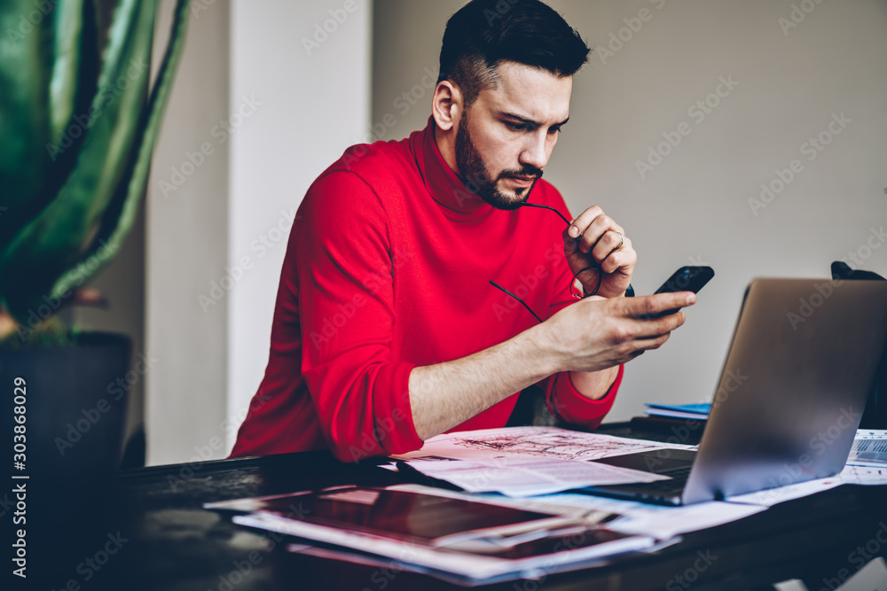 Poster pensive young man reading incoming notification on smartphone while working on freelnace at modern l