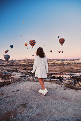 A tourist girl wearing white sweater on a mountain top enjoying wonderful view of balloons in Cappadocia