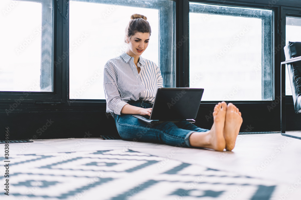 Poster smiling young woman keyboarding on laptop computer and updating app for remote work sitting on floor