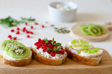 Open vegetarian sandwiches with vegetables, fruits and cream cheese on a wooden board on a white background. Kiwi and pomegranate in the background. Healthy breakfast concept. Copy space.