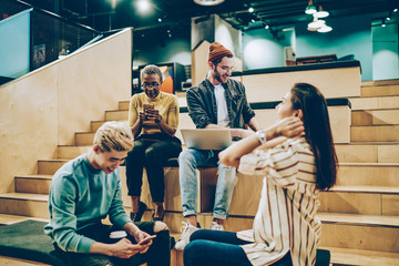 Positive multiracial colleagues communicating during free time on break in campus using modern gadgets, group of male of female hipsters spending time together share creative ideas and networking.