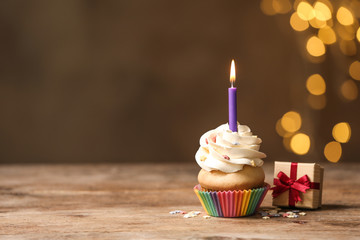 Birthday cupcake with candle and gift box on wooden table against blurred lights. Space for text