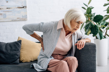 senior woman having back pain and sitting on sofa in apartment