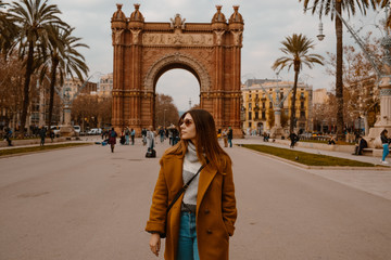 BARCELONA, SPAIN, January 17,2019.  Girl on the background the arch of Triumph in Barcelona. Cute young girl posing in autumn park. She is wearing mustard coat and glasses.  - obrazy, fototapety, plakaty