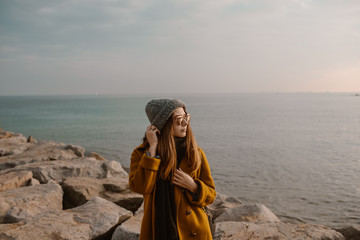 Girl portrait on the beach with the sea in the background. Stylish beautiful girl in mustard coat and glasses walking near the sea. 