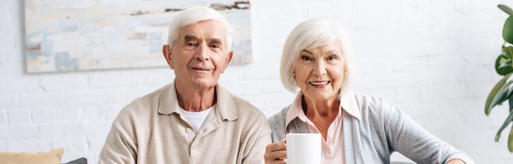 panoramic shot of husband and smiling wife holding cup and looking at camera  in apartment