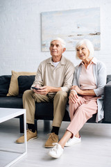 Obraz premium smiling husband and wife watching tv and sitting on sofa in apartment