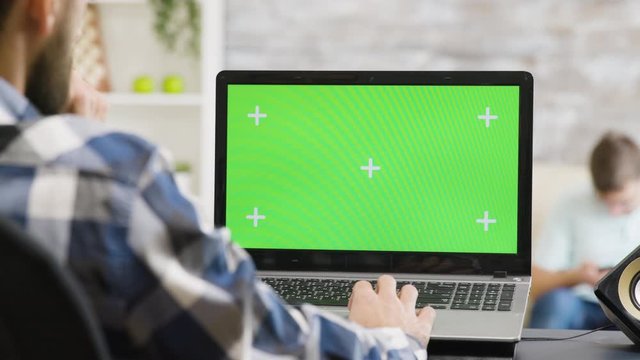 Over the shoulder shot of man working on laptop with green screen display in very bright living space