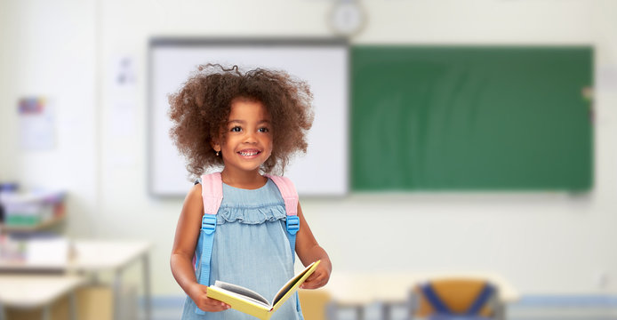 childhood, school and education concept - happy little african american girl with book and backpack over classroom background