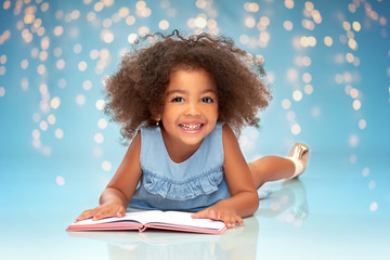 childhood, school and education concept - happy smiling little african american girl reading book over festive lights on blue background - obrazy, fototapety, plakaty