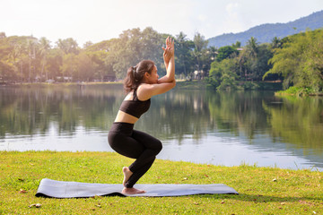 Fototapeta na wymiar Asian woman practicing yoga doing Eagle Pose pose on the mat beside a lake in outdoor park.