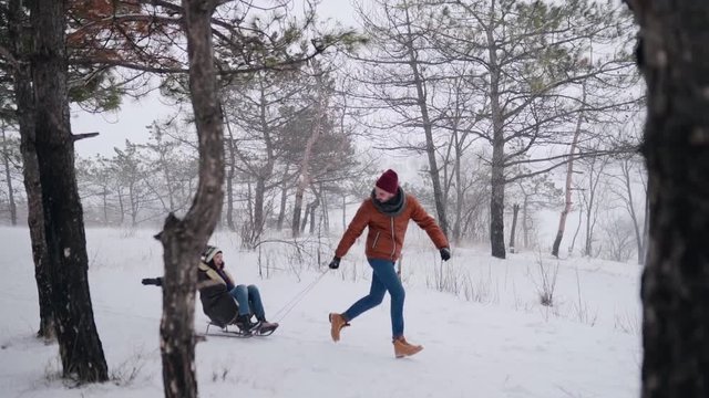 Lovely couple sledding on snowy winter day. Man pull sled with girlfriend on snowfall. Woman have fun and sledge outdoors with boyfriend. People sleigh ride and enjoy Christmas vacation. Slow motion.