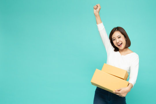 Happy Asian woman holding package parcel box and celebrating success with hands in the air isolated on light green background, Delivery courier and shipping service concept