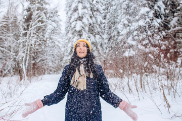 Portrait of a brunette woman in a yellow hat on a background of a winter forest. Smiling girl is playing with snow in the park.