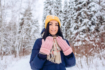 Portrait of a brunette woman in a yellow hat on a background of a winter forest. Smiling girl is playing with snow in the park.