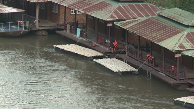 Several buildings docked on a flowing Kwai River, with wooden rafts tied onto them