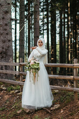 Beautiful elegant bride with hijab in lace wedding dress with long full skirt and long sleeves. She is holding a big bouquet of flowers. Outdoors, in the woods.