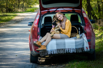 happy Caucasian girl sits in trunk of car, hugs a German shepherd dog and smiles, cheerful playful teen puppy looks at camera. Friendship of man and animal, travel, camping. Autumn forest. Copy space