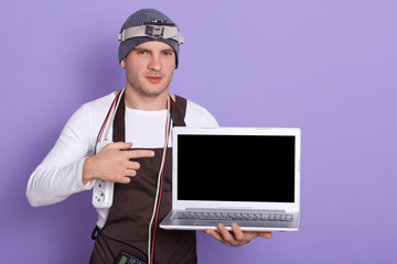 Young radioman wears white casual shirt, cap and apron, poins at blank screen of laptop over lilac background, radiotrician has extension around neck and other devices. Ecectronic engineering concept.
