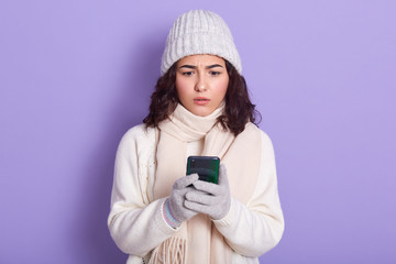 Adorable young girl looking concerned with text message on her smart phone, reads about bad news, has astonished facial expression, wears warm sweater, cap, scarf and gloves, isolated over lilac wall.