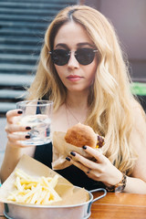 A young woman is eating hamburger, cheeseburger and french fries (chips), woman has yellow hair and she is enjoying with unhealthy food